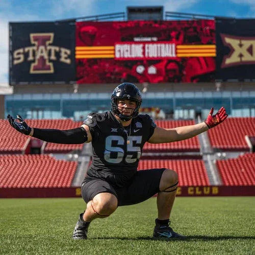 Football player in black uniform, number 65, crouching on field with stadium scoreboard behind